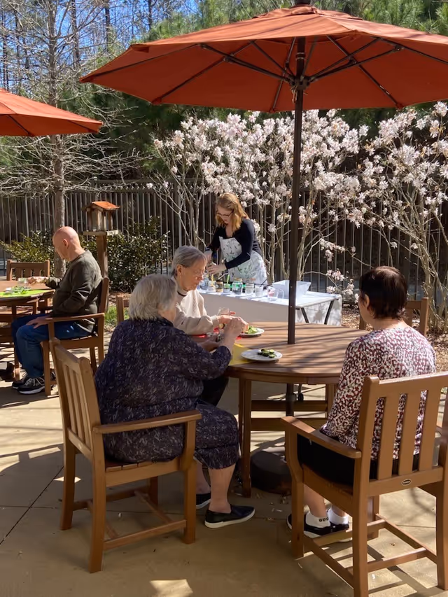 Several older adults sit at wooden patio tables under red umbrellas eating outdoors while a staff member arranges items on a nearby table with flowering trees behind them.