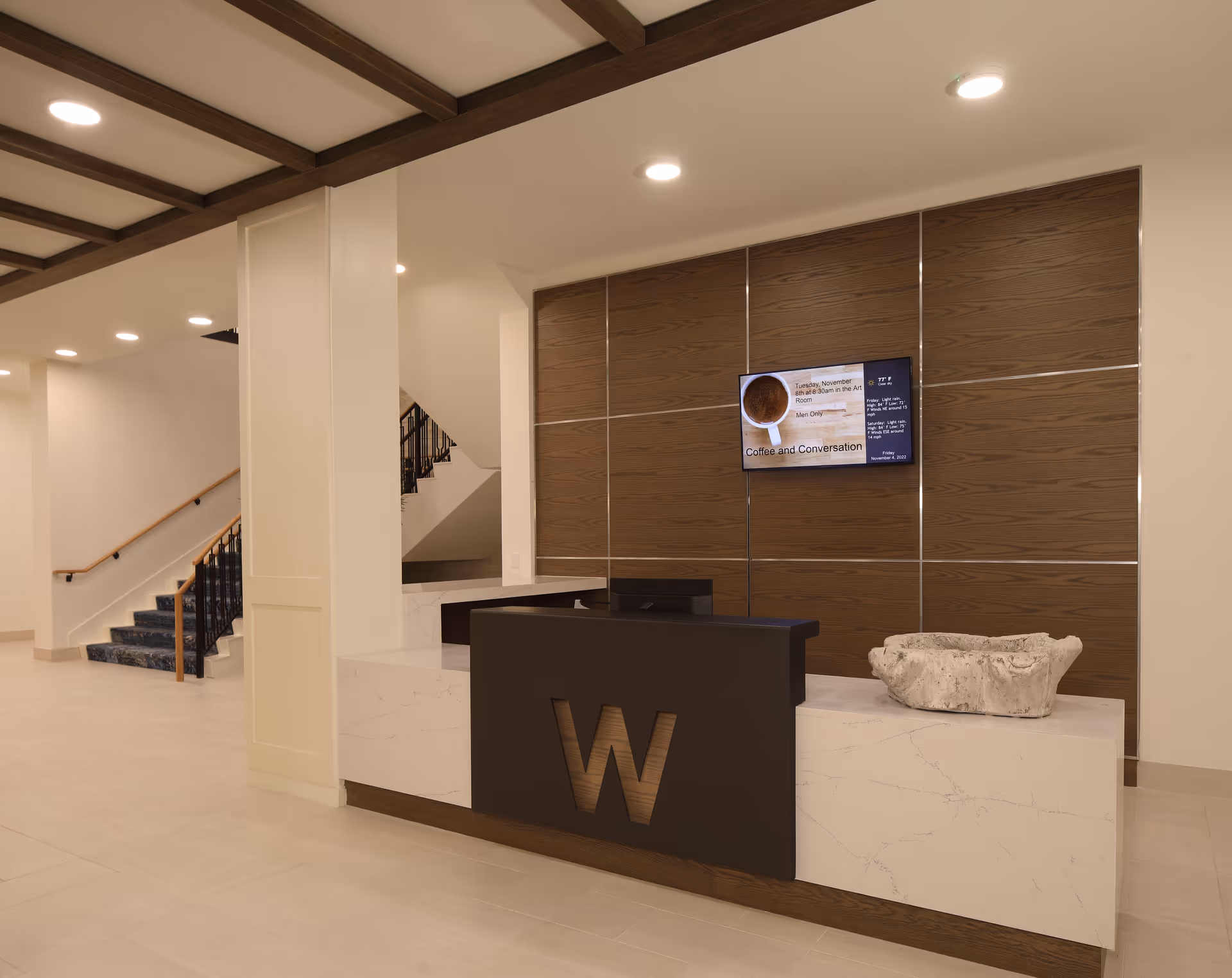 Reception area with a modern desk featuring a large letter W on the front, a stone decorative piece on the counter, a wooden paneled wall behind with a mounted digital screen displaying information, and a staircase with a carpeted runner and wooden handrails in the background.