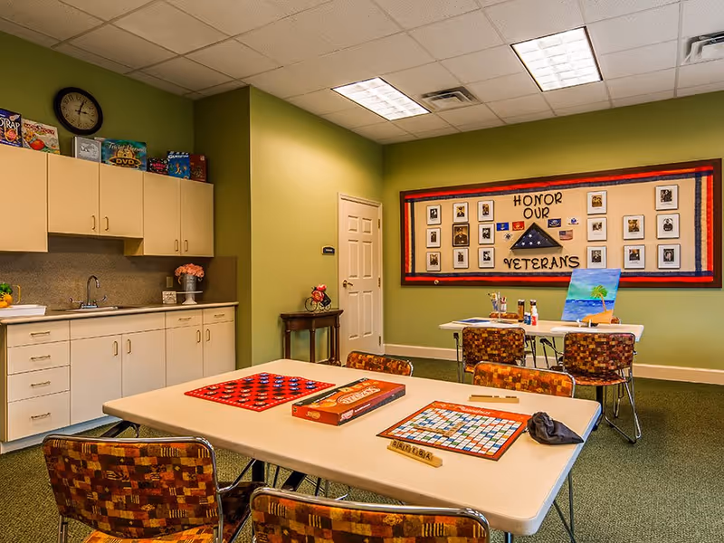 A community room with green walls featuring a kitchenette with cabinets, a sink, and a clock. Two tables with chairs are set up for games and painting activities. A bulletin board on the wall displays photos and a folded flag with the words 'Honor Our Veterans'.