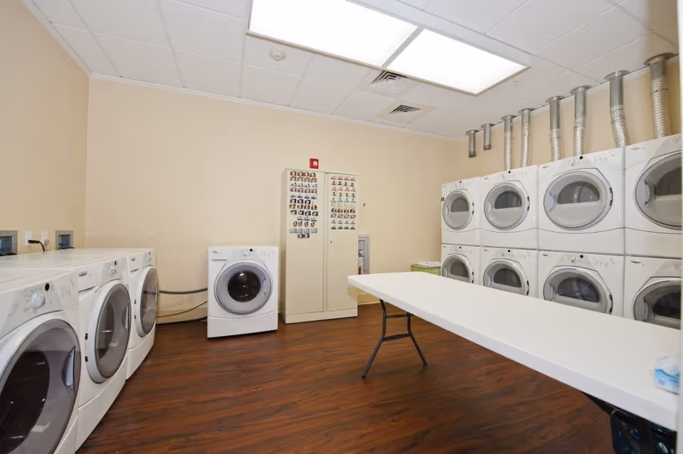 A laundry room with multiple white front-loading washing machines and dryers stacked on the right wall and lined up on the left wall. There is a white folding table in the center of the room and a beige cabinet with photos on it against the back wall. The floor is wood and the walls are light beige.