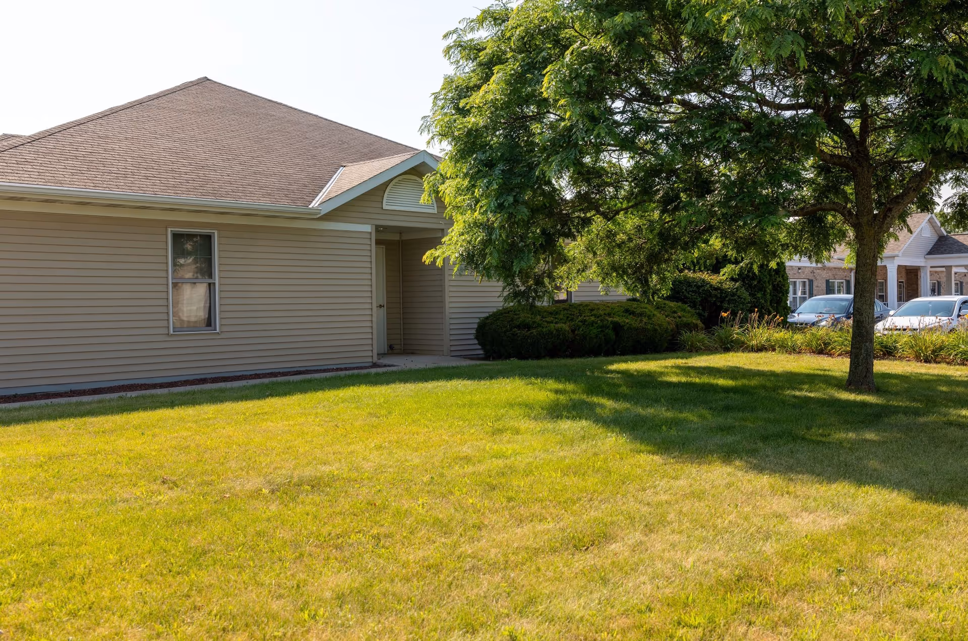 Single-story beige siding building with a grassy lawn, a tree in the foreground, and parked cars visible in the background.
