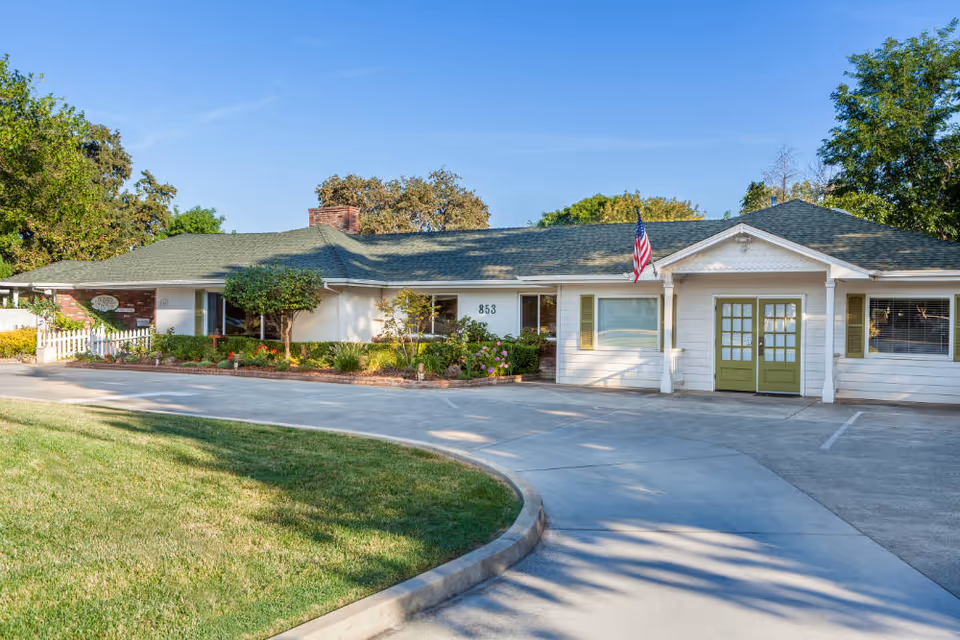 Single-story assisted living building front with a green roof, double green entry doors, an American flag, curved driveway and landscaped lawn.