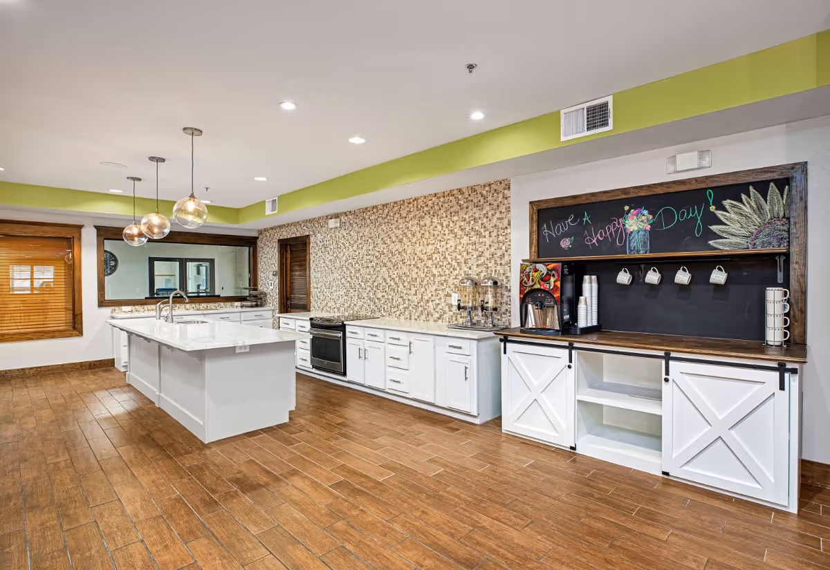 A modern kitchen area with a large white island countertop, three hanging pendant lights, white cabinets, a stove, and a mosaic tile backsplash. To the right, there is a coffee station with a chalkboard above it displaying colorful drawings and the message 'Have A Happy Day!'. The floor is wooden, and there is a large mirror on the wall opposite the island.