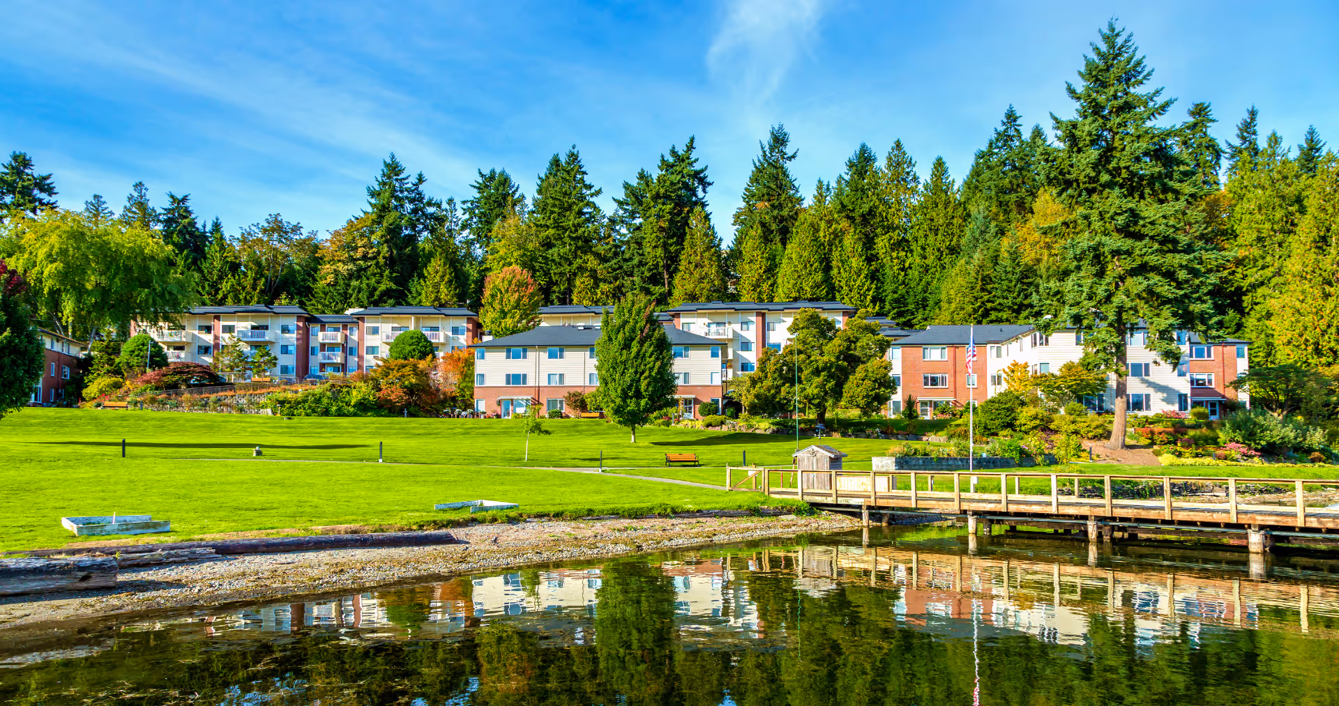 View of a senior living facility named Covenant Living at the Shores, showing multiple residential buildings surrounded by lush green trees and a well-maintained grassy area. In the foreground, there is a calm body of water with a wooden dock extending over it, reflecting the buildings and trees. The sky is clear with a few clouds.