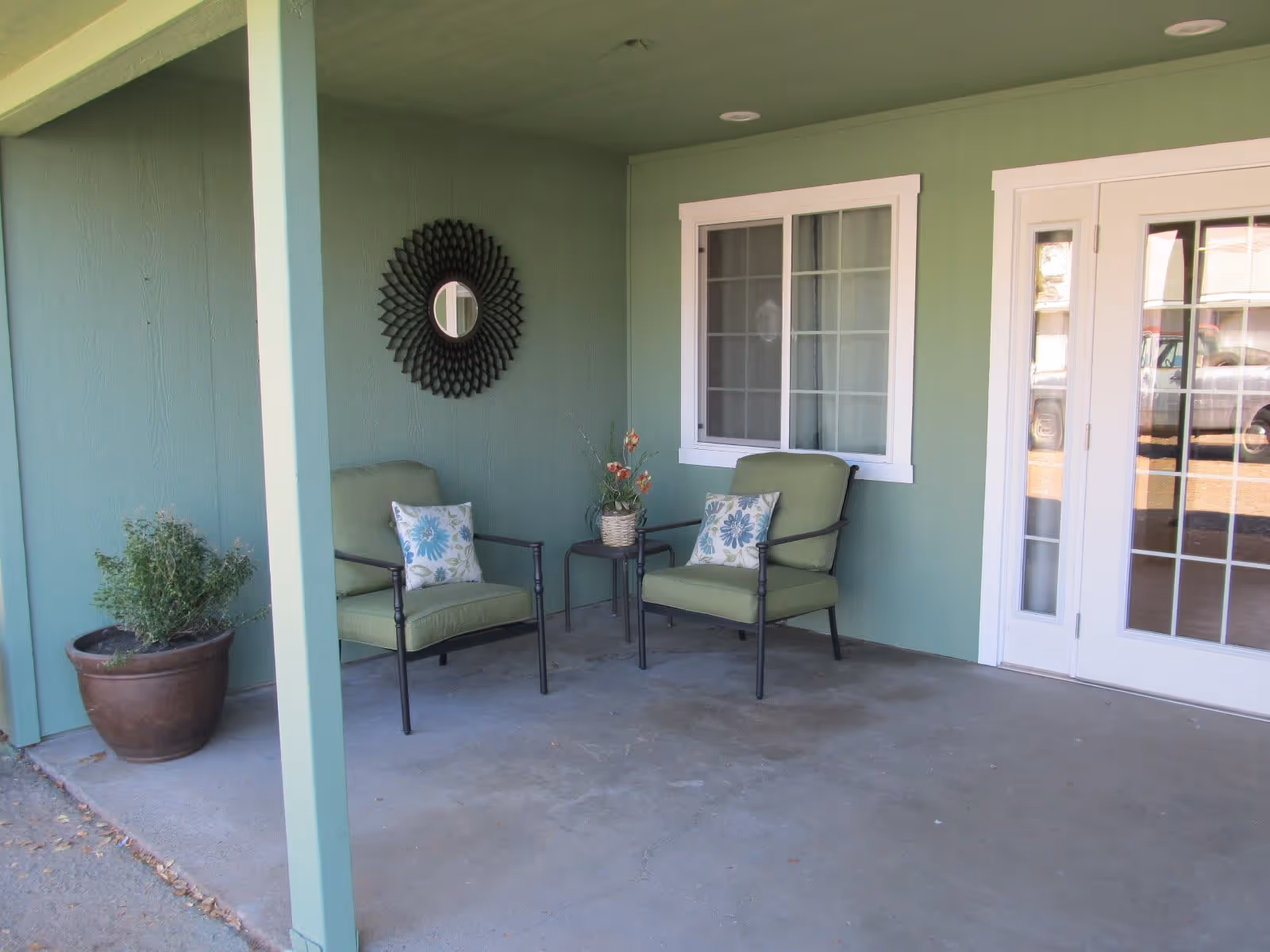 Covered outdoor patio area with two green cushioned chairs, each with a floral pillow, a small side table with a flower arrangement, a large potted plant, a decorative round mirror on the green wall, a window with white trim, and white double doors with glass panes.