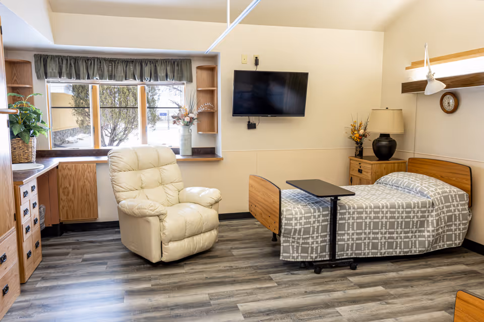 A senior living facility bedroom with a single bed covered in a gray and white patterned blanket, a beige recliner chair, a wall-mounted flat screen TV, a wooden nightstand with a lamp and flower arrangement, a clock on the wall, and a window with green valance curtains letting in natural light.
