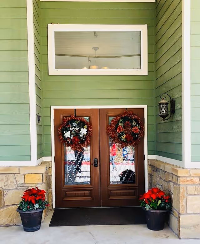 Double wooden front doors decorated with festive wreaths, flanked by two black pots with red poinsettia plants. The exterior walls are green with stone accents and a lantern-style light fixture on the right side.