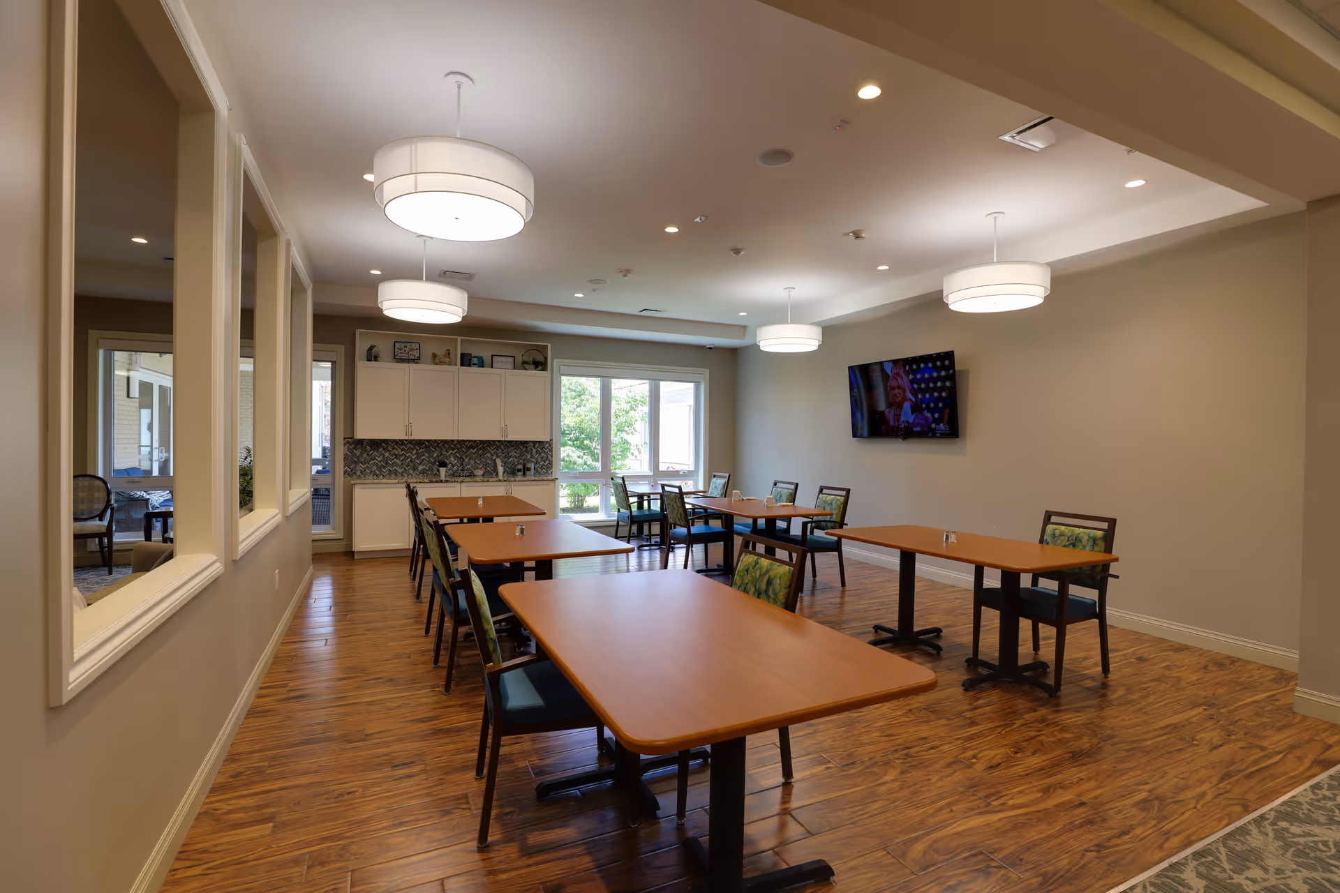 Well-lit communal dining room with wooden tables and chairs, pendant lights, cabinetry and a wall-mounted TV.
