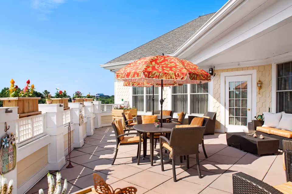 Outdoor patio area with a round table and six wicker chairs under a red and yellow floral umbrella. There are additional wicker seating arrangements with cushions, potted plants on the railing, and a clear blue sky in the background.