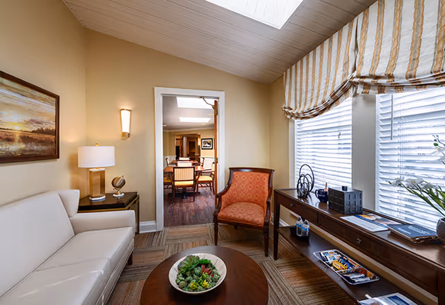A cozy living room area with a white sofa on the left, a wooden coffee table with a bowl of succulents in the center, and an orange upholstered armchair near a window with striped Roman shades. A wooden console table under the window holds decorative items, books, and magazines. The room has beige walls, a framed landscape painting, a table lamp, and a wall sconce. Through an open doorway, a dining area with a wooden table and chairs is visible.