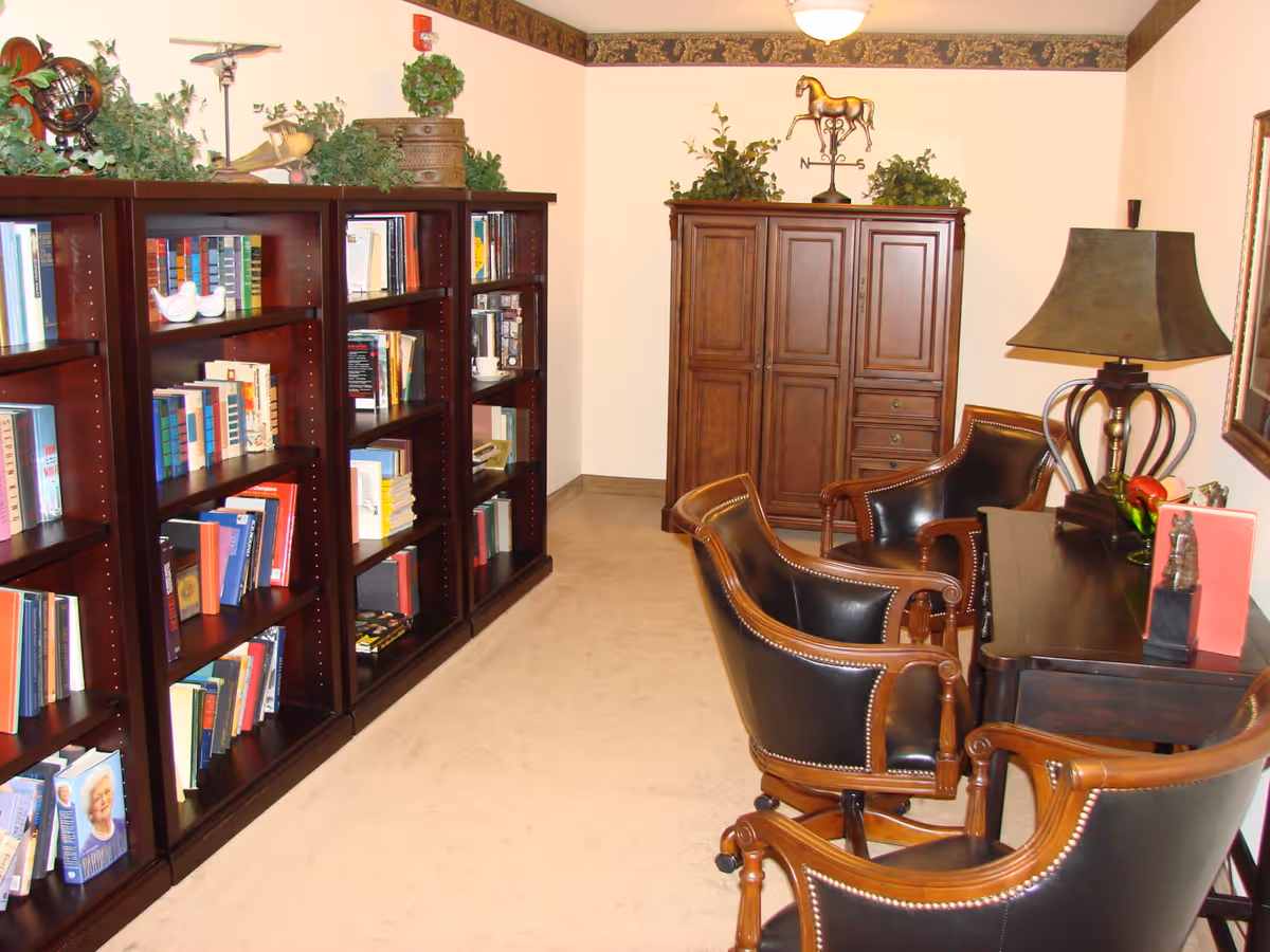 A cozy reading room with dark wooden bookshelves filled with books, three leather and wood armchairs around a dark wooden desk with a lamp, and a wooden cabinet against the back wall decorated with plants and a horse figurine on top.