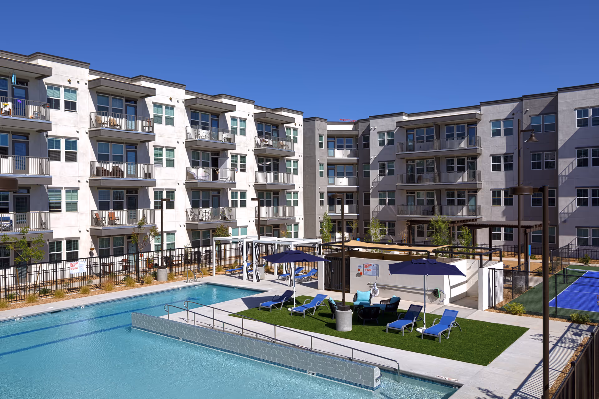 Outdoor view of a senior living facility named Mera Rhodes Ranch featuring a large swimming pool with clear blue water, surrounded by lounge chairs and umbrellas on a grassy area. The background shows a multi-story residential building with balconies and windows under a clear blue sky.