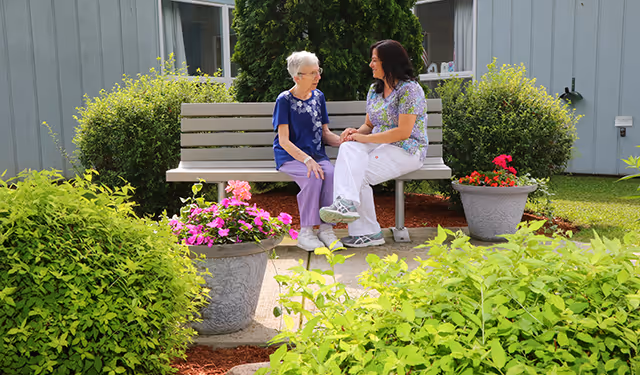 An elderly woman and a caregiver sitting on a bench outdoors in a garden area, surrounded by green bushes and colorful flowers, engaged in a friendly conversation.