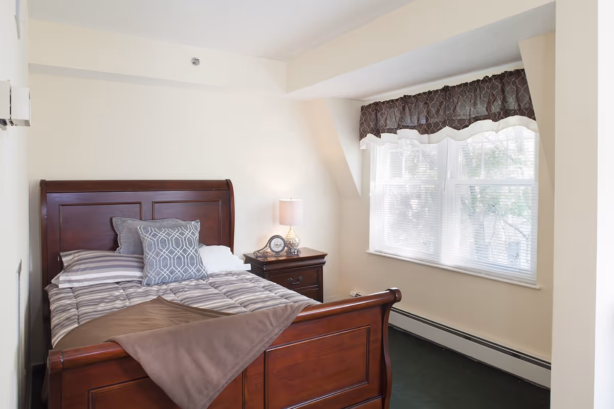 Bright bedroom featuring a wooden sleigh bed with striped bedding, a nightstand with a lamp and clock, and a large window with blinds.