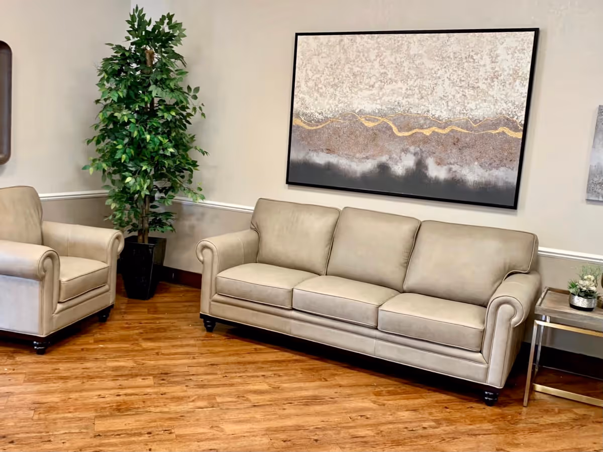 A cozy seating area with a beige leather sofa and matching armchair on a wooden floor. There is a tall green potted plant in the corner and a side table with a small plant on it. A large abstract painting with neutral tones and gold accents hangs on the wall above the sofa.