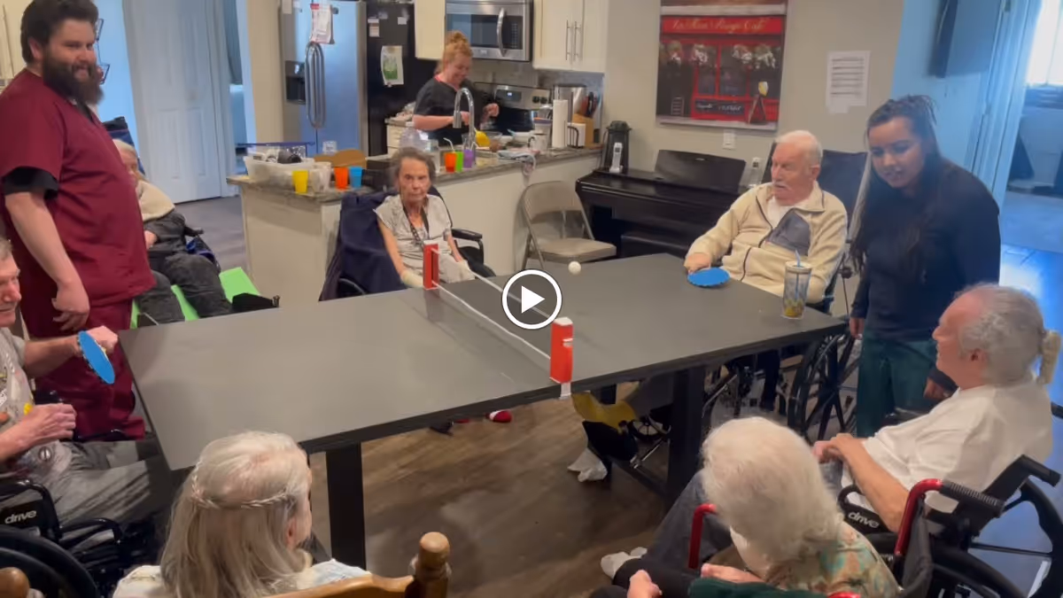 A group of elderly people, some in wheelchairs, playing table tennis indoors around a small ping pong table. Two caregivers are present, one standing near the table and another in the kitchen area. The setting appears to be a common area with a kitchen in the background.