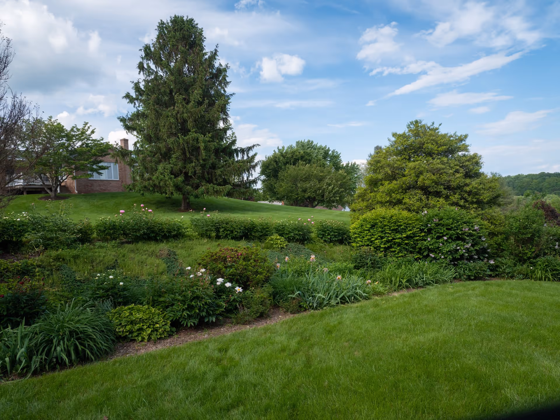 A landscaped garden area with lush green grass, various bushes, flowering plants, and several trees under a partly cloudy blue sky. A brick building with large windows is partially visible in the background on a small hill.