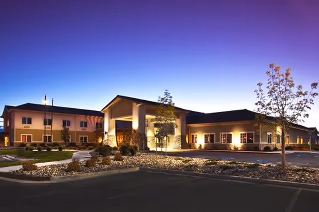 Front exterior of a skilled nursing and rehabilitation facility at dusk with an illuminated entrance canopy, landscaped grounds, and a parking area.