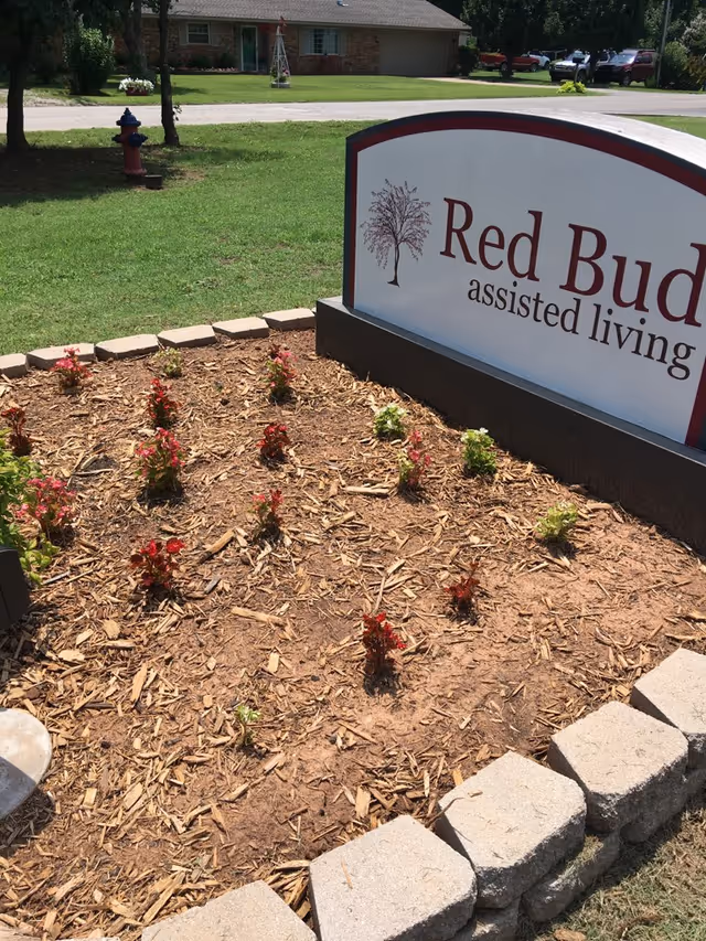 A landscaped garden bed with small flowering plants and mulch in front of a sign that reads 'Red Bud assisted living'. In the background, there is a grassy lawn, a fire hydrant, a residential house, and several parked vehicles.