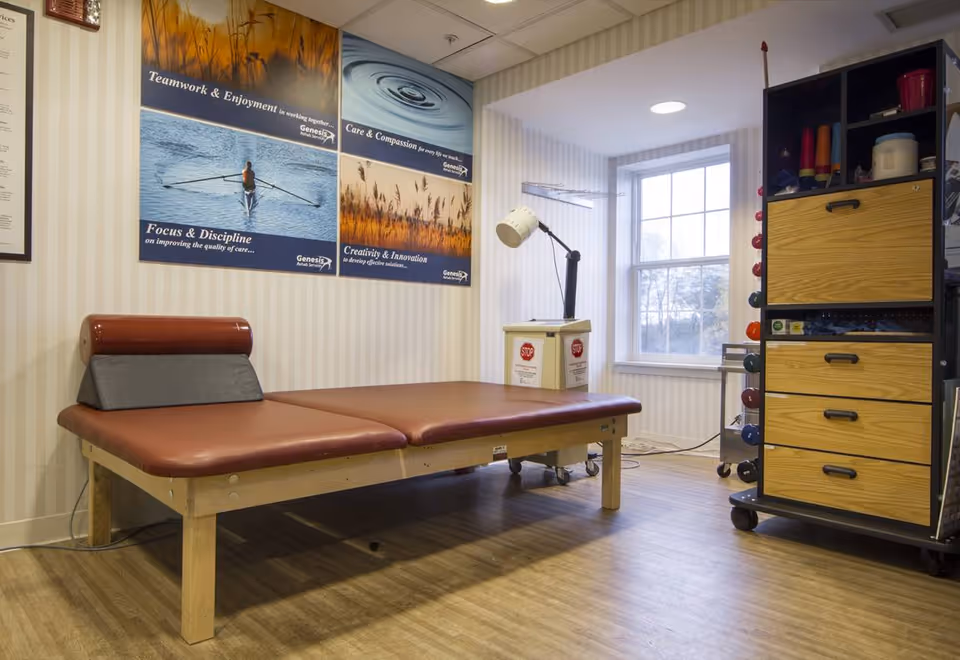 A therapy room with a padded treatment table, adjustable lamp, storage cabinet with weights, and motivational posters on the wall.