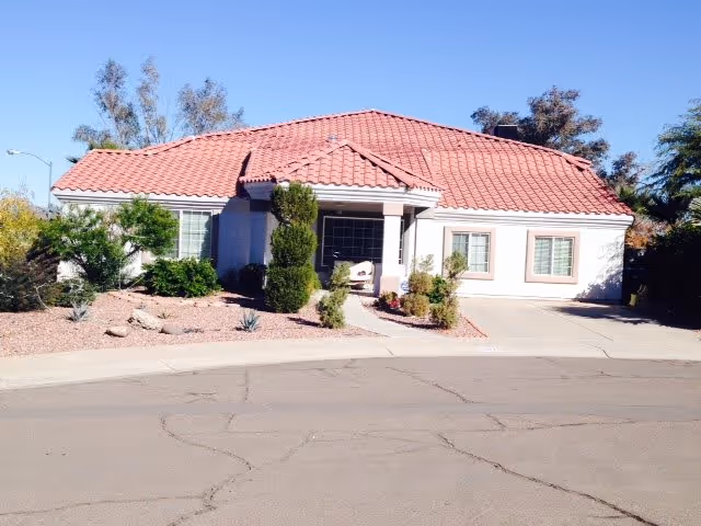 Single-story building with a red tiled roof and light-colored exterior walls, surrounded by desert landscaping with shrubs and small trees, situated on a paved street under a clear blue sky.