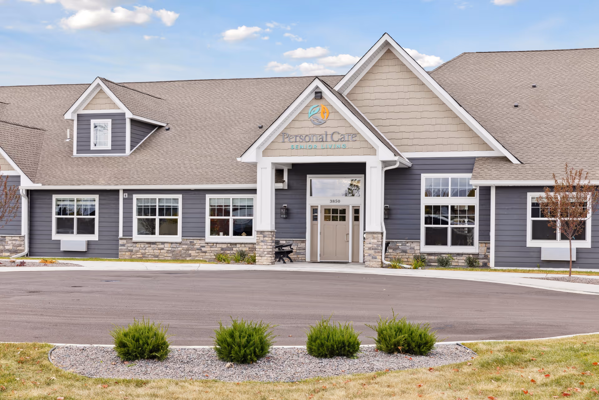 Exterior front view of a single-story senior living facility building with gray siding, stone accents, multiple windows, and a peaked roof. The entrance has a covered porch with white columns and a sign above reading 'Personal Care Senior Living'. There are small bushes and a landscaped area in front of the building under a partly cloudy sky.