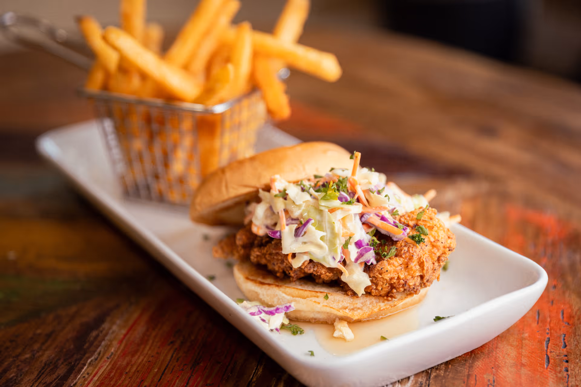 A close-up of a fried chicken sandwich topped with coleslaw on a white rectangular plate, served with a small metal basket of French fries on a wooden table.