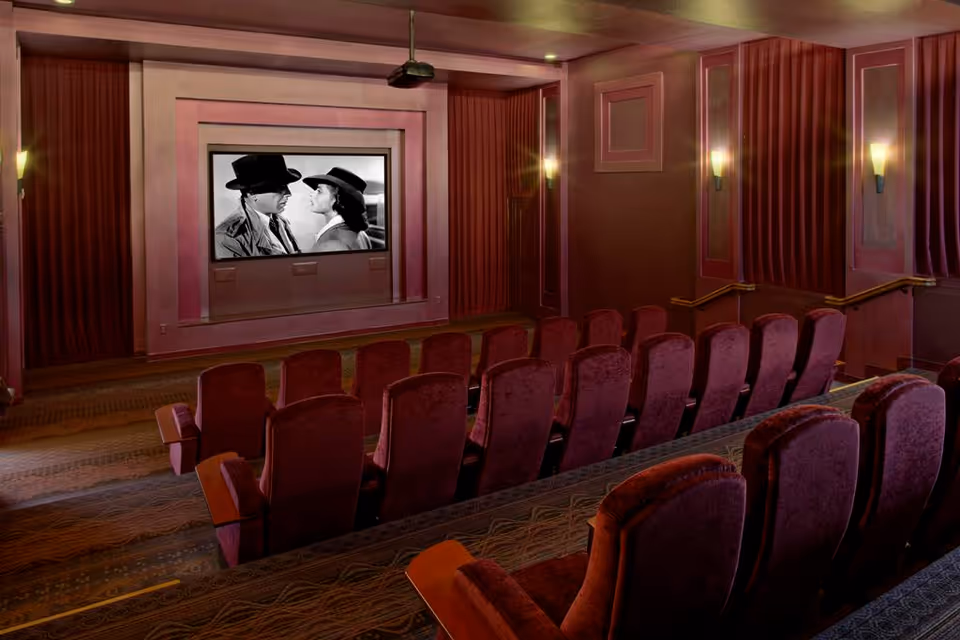 Small movie theater screening room with rows of red upholstered seats facing a screen showing a black-and-white film.