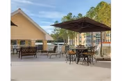 Outdoor patio area with several tables and chairs, including a table with a large umbrella. There is a small building or shed on the left side and trees in the background under a clear blue sky.