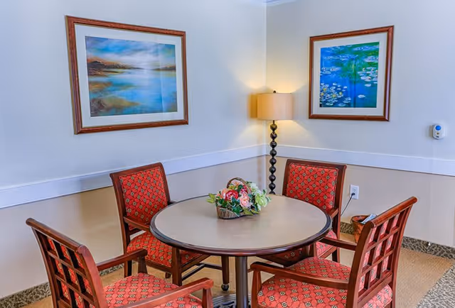 A small round table with a floral centerpiece surrounded by four wooden chairs with red patterned cushions in a corner of a room. Two framed paintings hang on the walls above the table, and a tall floor lamp with a beige shade is positioned in the corner.