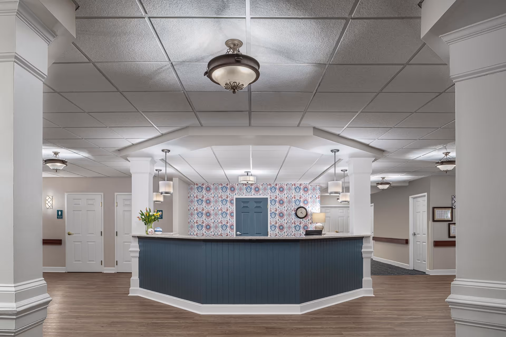 A reception desk in a senior living lobby with a blue paneled front, patterned wallpaper behind it, pendant lights, and surrounding hallways.