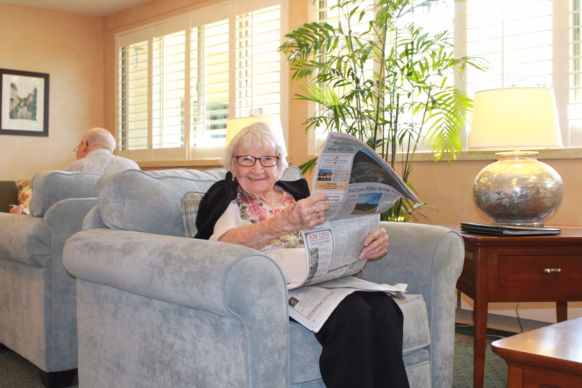 An elderly woman with glasses sitting on a light blue armchair in a well-lit living room, smiling and reading a newspaper. Behind her, an elderly man is seated on another armchair. The room has large windows with white shutters, a green plant, a wooden side table with a lamp, and a framed picture on the wall.