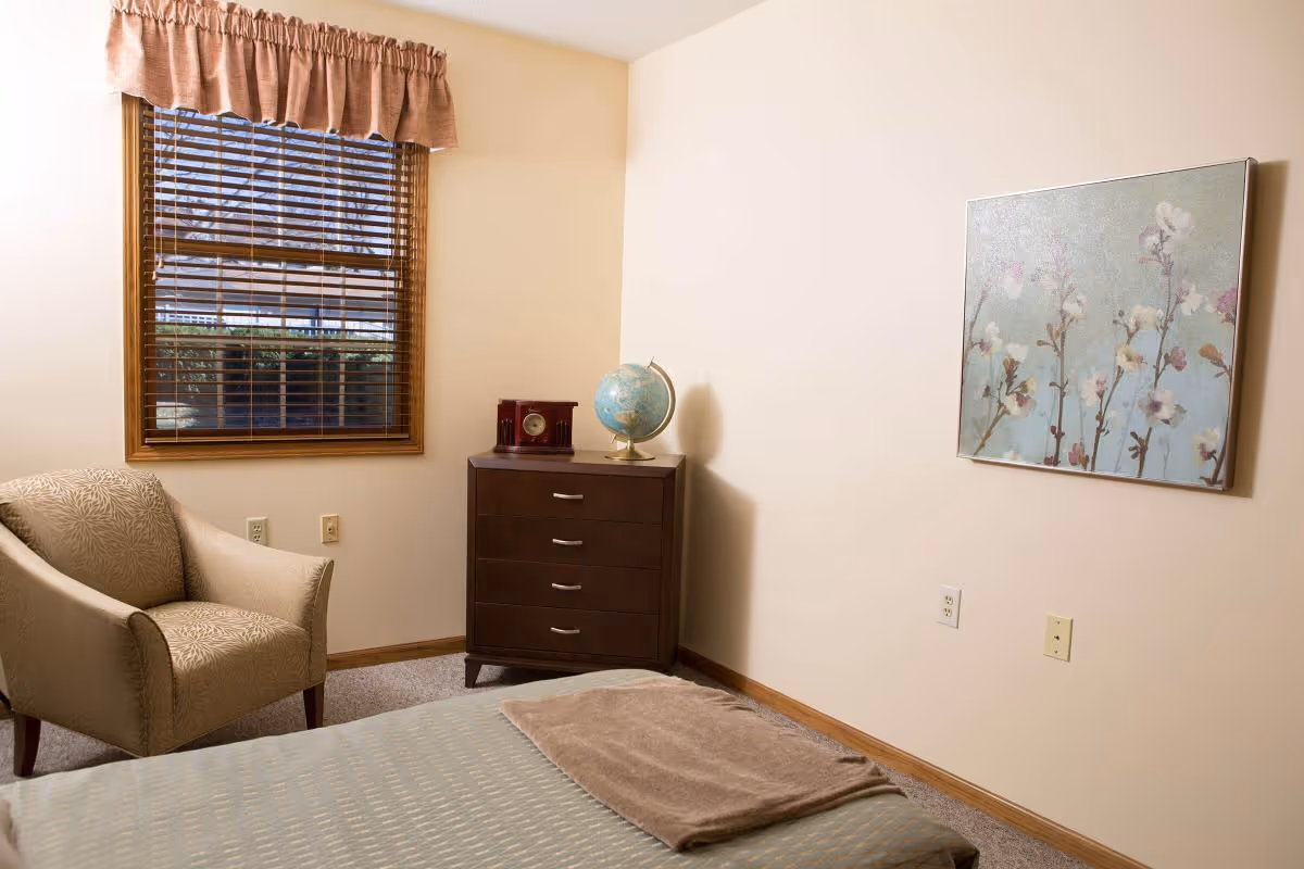 A cozy bedroom with a beige armchair, a wooden dresser topped with a globe and a clock, a bed with a folded blanket, a window with wooden blinds and a valance, and a floral painting on the wall.