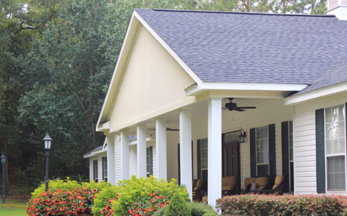 Front porch of a light-colored assisted living building with white columns, outdoor seating, and landscaped shrubs.