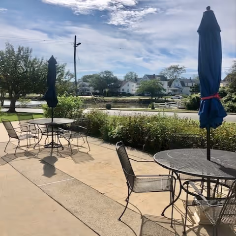 Outdoor patio area with metal tables and chairs, each table having a closed blue umbrella. There is a hedge bordering the patio and a view of a street, trees, and houses across the street under a partly cloudy sky.