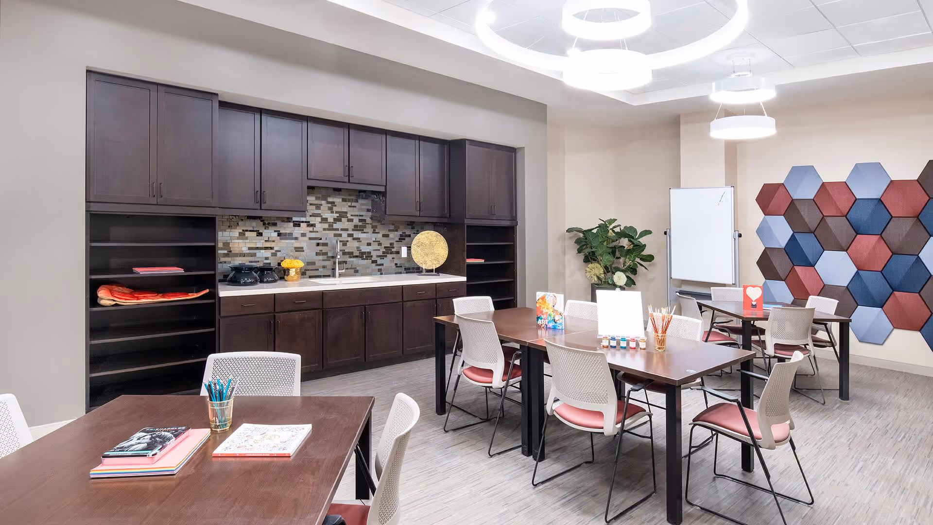 A bright and modern activity room with dark wooden cabinets and a mosaic tile backsplash along one wall. The room features several tables with white chairs that have pink cushions. On the tables are art supplies including colored pencils, paint bottles, and paper. A large plant and a whiteboard are visible in the corner, and a colorful hexagonal wall decoration adds visual interest. The ceiling has contemporary circular light fixtures.