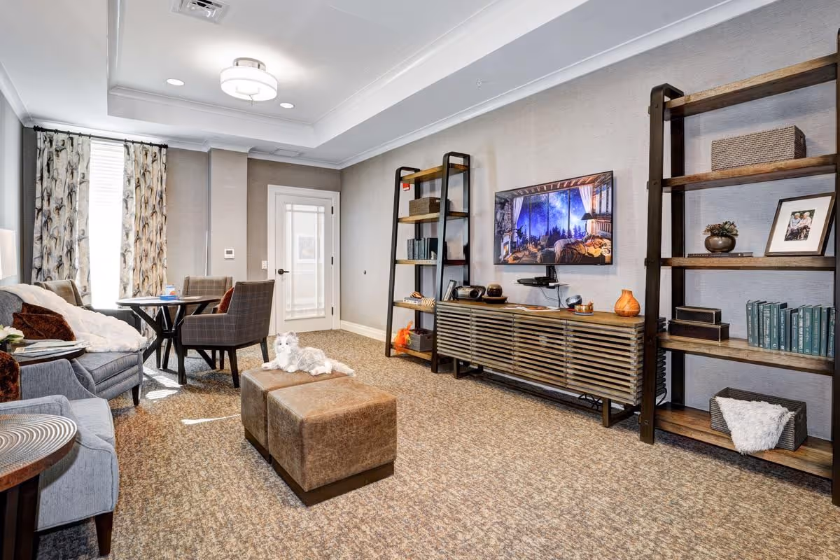 Well-appointed living room with seating, ottomans, a wall-mounted TV above a media console, open shelving, and a small dining table near a window.
