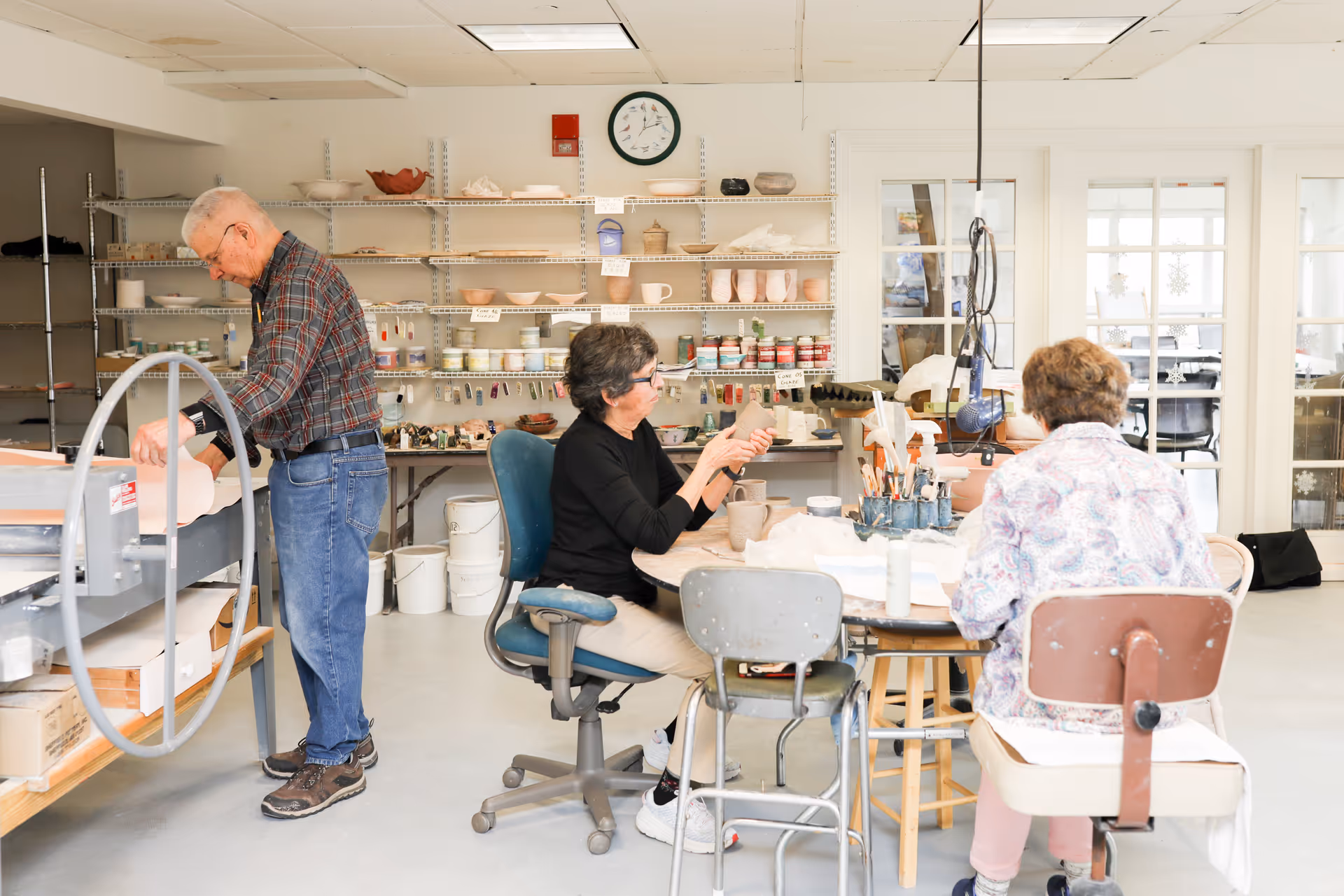 Three elderly individuals engaged in pottery activities in a bright, organized art studio. One man is standing near a pottery wheel, while two women are seated at a round table working on clay pieces. Shelves in the background hold various pottery supplies and finished pieces.