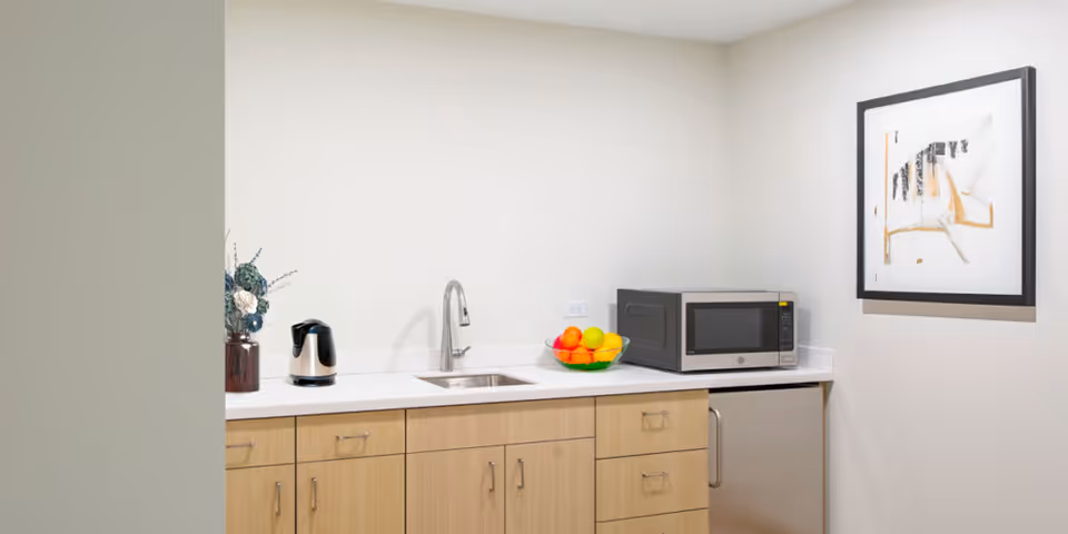 A small kitchenette area with light wood cabinets, a stainless steel sink with a modern faucet, a microwave, a small refrigerator, a bowl of assorted fruit, a kettle, and a decorative vase with flowers. A framed abstract artwork hangs on the wall.