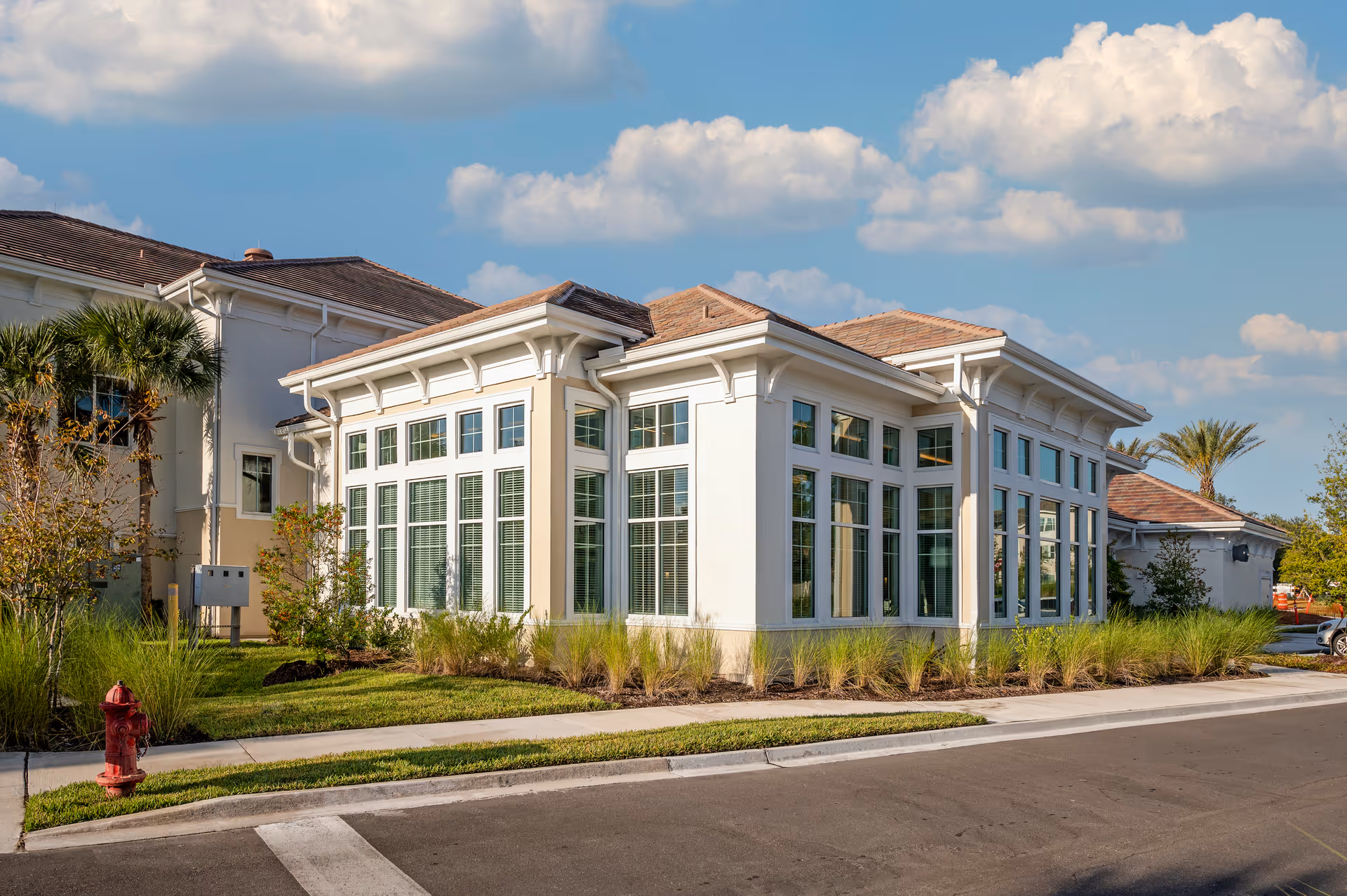 Exterior view of a modern senior living facility building with large windows, surrounded by landscaped greenery including palm trees and grass, under a partly cloudy blue sky.