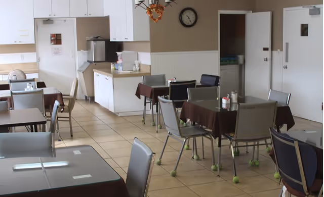 Interior view of a dining room with several tables covered with brown tablecloths and surrounded by chairs with tennis balls on the legs. There is a kitchenette area with white cabinets, a microwave, and a water dispenser. A clock is mounted on the wall above the kitchenette, and there are doors visible in the background.