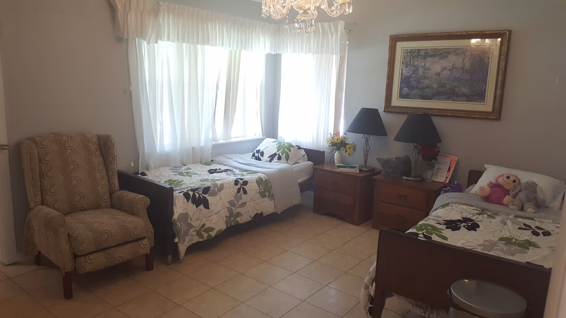 A tidy shared bedroom with two single beds, bedside tables and lamps, an armchair, and a large curtained window.