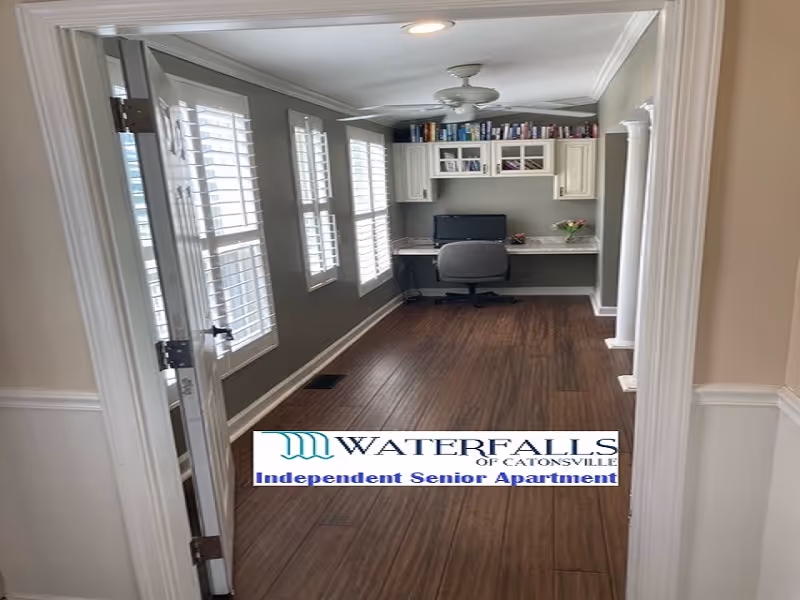 View through a doorway into a bright home office/study with a built-in desk, chair, bookshelves and wood flooring.