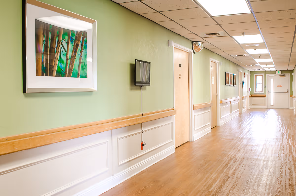 Well-lit interior hallway in a senior living facility with green walls, framed artwork, handrails and multiple doors along a wood floor.