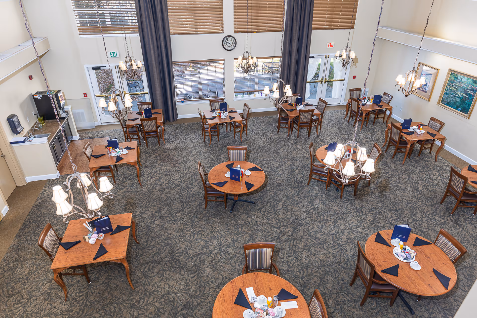 A spacious dining room with multiple wooden tables and chairs arranged neatly. Each table has black napkins, menus, and some have plates and cups. The room features large windows with blinds, chandeliers hanging from the ceiling, and framed artwork on the walls. There are two exit doors visible, and a clock is mounted on the wall above the windows.