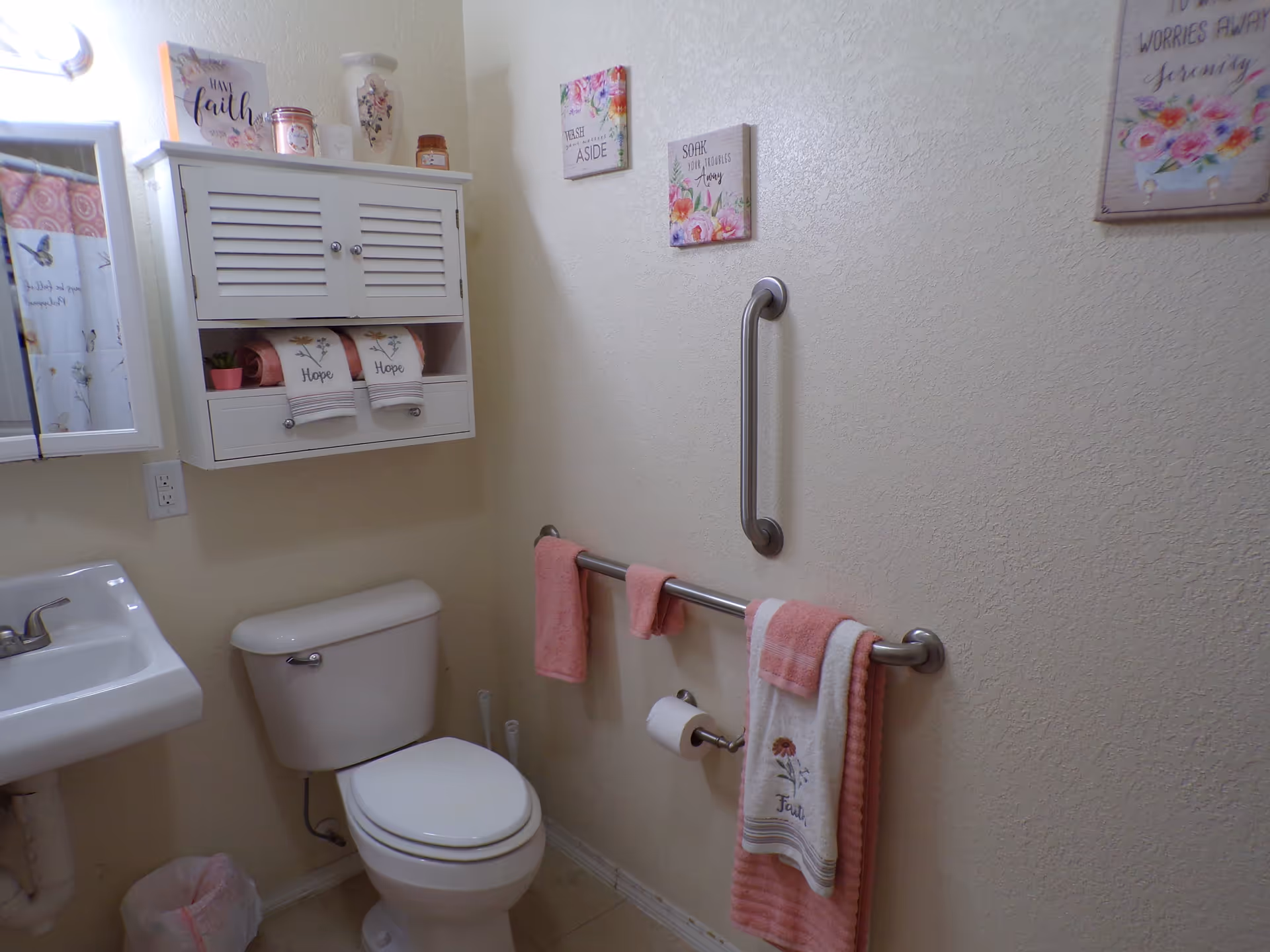 A small bathroom with a white toilet and sink. Above the toilet is a white wall cabinet with two doors, two drawers, and an open shelf holding pink towels and decorative items. The wall has three floral-themed framed signs with inspirational messages. There are two stainless steel grab bars on the wall, one horizontal with pink towels hanging on it and a toilet paper holder, and one vertical. The bathroom has light-colored walls and floor tiles.