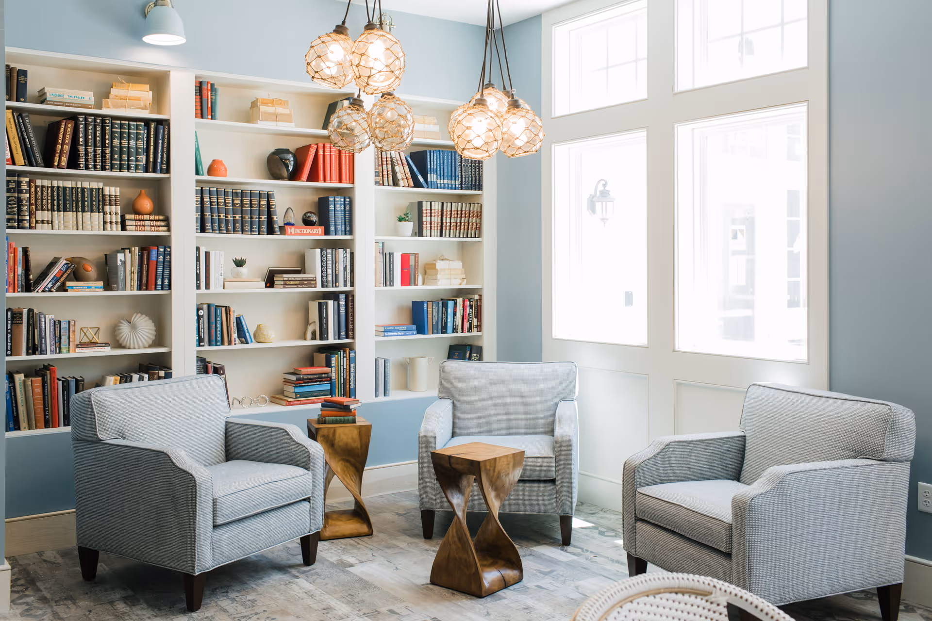 A cozy sitting area with three light gray armchairs arranged around two small wooden side tables. Behind the chairs is a large built-in bookshelf filled with books and decorative items. The room has light blue walls and large windows letting in natural light. Three pendant lights with woven shades hang from the ceiling.