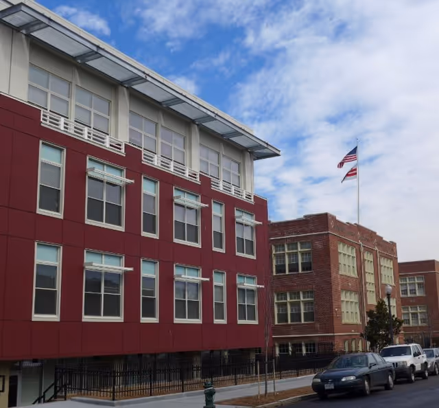 Red brick and red panel exterior of Allen House Senior Apartments with parked cars along street.