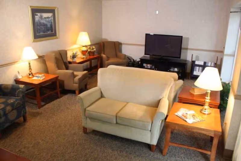 A cozy living room area with a beige loveseat in the center, two beige armchairs against the wall, a blue patterned armchair on the left, wooden side tables with lamps, a flat-screen TV on a black TV stand, and a framed picture on the wall.