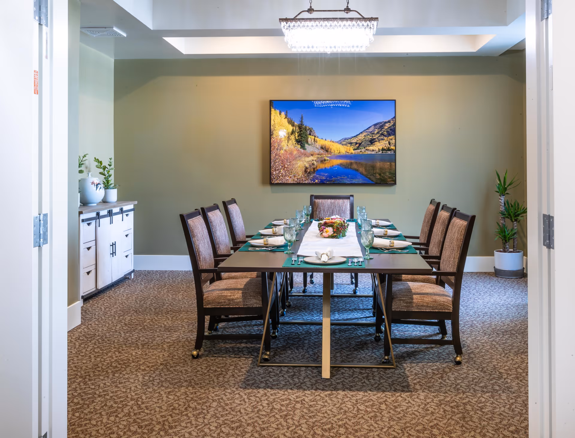 Dining room with a long table set for eight, upholstered chairs, a landscape picture on the wall, and a sideboard with plants.